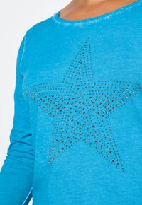 Türkisfarbiges Langarmshirt mit einem großen Sternmuster aus silbernen Nieten. Das Shirt hat einen runden Ausschnitt und eine weiche Textur.