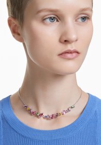 Multicolored gem necklace with pink, purple, and green stones, set in a silver chain, worn against a light blue ribbed top.