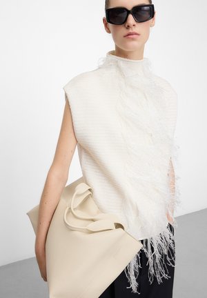 Frau trägt übergroße schwarze Sonnenbrille und ein ärmelloses weißes strukturiertes Oberteil mit Fransen, hält eine große beige Handtasche.