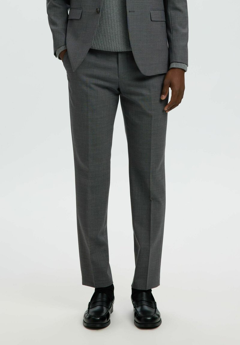 Homme portant un pantalon de costume gris, un pull gris, une veste de costume grise et des mocassins en cuir noirs, debout avec les mains détendues.