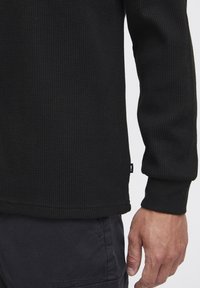 Black waffle-knit long-sleeve shirt with a round neck and ribbed cuffs, featuring a small, discreet logo tag on the side.