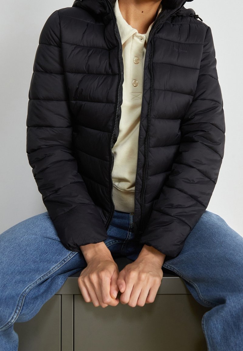 Czarna kurtka puchowa z zapięciem na zamek i kapturem, z pikowanymi elementami, w połączeniu z jasnym polo i niebieskimi dżinsami.