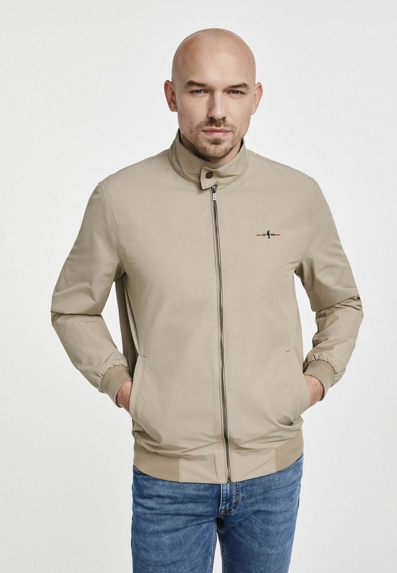 Jasno beżowa kurtka z wysokim kołnierzem, przednim zamkiem błyskawicznym i elastycznymi mankietami. Zawiera haftowane logo i dwie boczne kieszenie. Nosi się ją na niebieskich dżinsach.