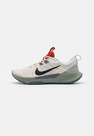 Chaussure de running Nike beige clair et grise avec swoosh noir, languette orange et semelle épaisse verte sur fond blanc.
