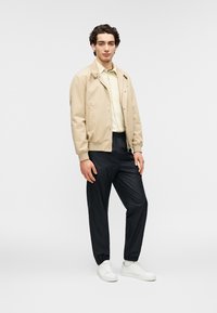 Μπεζ μπουφάν bomber πάνω από ανοιχτόχρωμο πουκάμισο, συνδυασμένο με μαύρο παντελόνι με κωνικό τελείωμα και λευκά αθλητικά παπούτσια. Λεπτά υφάσματα, casual εφαρμογή και μινιμαλιστικός σχεδιασμός.