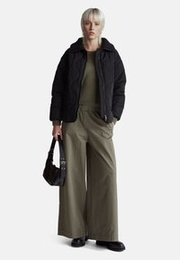 Černá prošívaná bunda s vysokým límcem, olivově zelený pletený svetr, volné olivově zelené kalhoty se širšími nohavicemi a černá kabelka přes rameno.