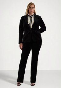 Svart sammetsdräkt med en figursydd kavaj och byxor med raka ben. Kavajen har en enkel knapp, medan en vit blus med volanger finns under.