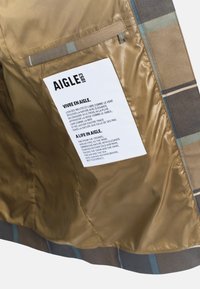 Doublure de veste à carreaux marron avec une texture brillante. Comprend une étiquette avec du texte, une poche et des accents rayés en bleu et beige.
