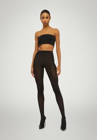 Leggings neri trasparenti con un design a vita alta, indossati con un top bandeau nero e scarpe con tacco alto nere. Texture liscia, design aderente.