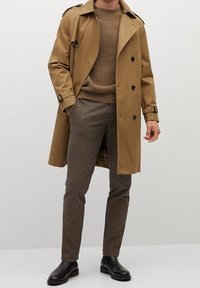Trench beige con botones de doble pecho, suéter de punto marrón y pantalones con estampado de pata de gallo. Botines negros completan el look.
