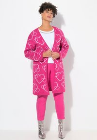 Horkorůžový kardigan s bílými vzory srdcí, navlečený přes bílý top a spárovaný s odpovídajícími růžovými kalhotami a lesklými stříbrnými botami.