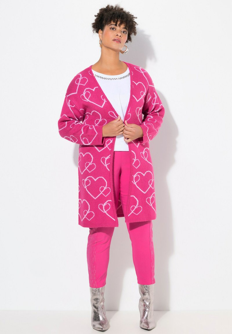 Horkorůžový kardigan s bílými vzory srdcí, navlečený přes bílý top a spárovaný s odpovídajícími růžovými kalhotami a lesklými stříbrnými botami.