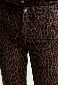Jean en denim imprimé léopard marron et noir, avec un design classique à cinq poches et un bouton à fermoir en métal. Tissu texturé avec un motif distinct.