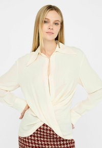 Blusa color crema con una texture liscia, ampio colletto e un design a spirale frontale. Maniche lunghe e vestibilità comoda.