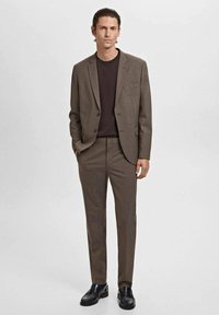 Man standing wearing a brown suit with matching trousers, black loafers, and a black crew neck shirt, hands in pockets against a plain background.