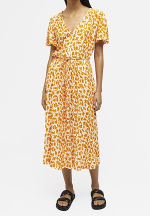 Kvinde iført en orange og hvid mønstret midikjole med flagrende ærmer, V-hals, snoretræk i taljen og sorte slip-on sandaler.