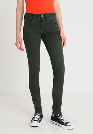 Osoba ubrana w ciemnozielone, dopasowane jeansy, czarne sneakersy za kostkę z białymi sznurowadłami oraz jaskrawo pomarańczowy, bezrękawnik, stojąca na tle białego tła.