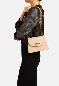 Borsa a mano in pelle beige con chiusura a pattina arrotondata, accessori in oro e tracolla regolabile, indossata su un abbigliamento nero con uno sfondo neutro.
