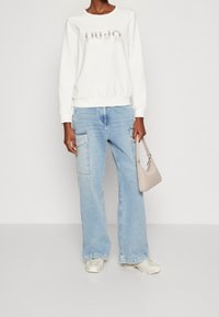 Osoba v bílém svetru s logem "Liu Jo", světlých modrých širokých kalhotách typu cargo, bílých teniskách a držící b�éžovou kabelku.