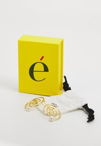 Zlatne spiralne naušnice s bisernim naglascima, predstavljene u svijetlu žutu kutiju s logom. Uključena je bijela torbica na vezivanje.