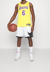 Žlutý basketbalový dres s fialovým nápisem "Lakers" a číslem "6", doplněný bílými šortkami s černým logem Nike a bílými teniskami.