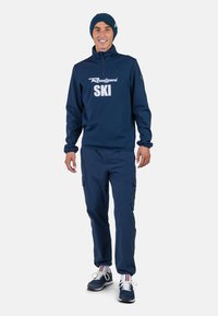 Pullover da sci in navy con stampa bianca "SKI", abbinato a pantaloni cargo navy e sneakers blu. Look completo con un berretto abbinato.
