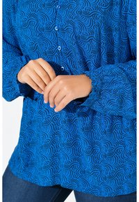 Blaue Bluse mit Muster, die spiralförmige schwarze Designs zeigt, mit Knopfverschluss und gerafften Ärmeln, aus leichtem Stoff gefertigt.