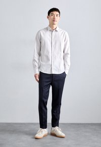Camisa de botones a rayas blanca y gris claro, pantalones navy ajustados y zapatillas ligeras con suela de goma. El modelo se encuentra de pie contra un fondo liso.