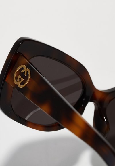 Lunettes de soleil en écaille de tortue avec des verres sombres, affichant une finition brillante. La monture présente un logo double G doré sur le côté pour le branding.