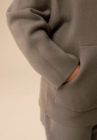 Grå stickad sweatshirt med texturerad tyg, sänkt axelsöm och en framficka med en synlig hand. Mjuk och avslappnad design.