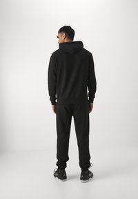 Sort hættetrøje og jogger-sæt, lavet af blødt stof, med hætte, elastiske manchetter og strækbart taljebånd. Båret med sorte sneakers.