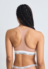 Világos rózsaszín sportmelltartó, versenyhátsó kialakítással, alján a "Calvin Klein" felirattal ellátott fehér elasztikus pánttal. Sima felület.