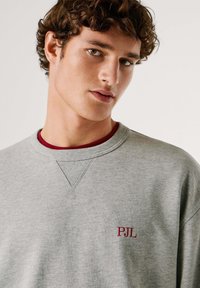 Felpa grigia con scollo a costine, caratterizzata da un piccolo logo rosso "PJL" sul davanti. Tessuto morbido con vestibilità rilassata e dettagli testurizzati.