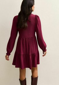 Bordowa sukienka z bufiastymi rękawami, z warstwową spódnicą i fakturowanym materiałem. Góra sukienki jest dopasowana, a rękawy mają elastyczne mankiety. Brązowe kozaki do kolan.