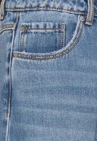 Světle modré džíny z denimu s malou zadní kapsou s třepenými okraji, zvýrazněné žlutým prošíváním a kovovými doplňky.