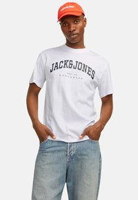 Hvid bomulds T-shirt med fed navy tekst "JACK & JONES" og "EST. 90 WORLDWIDE." Matchet med lyseblå denim jeans og en orange kasket.