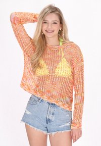Top trasparente a maniche lunghe in maglia con colori vivaci arancioni e gialli, indossato sopra un bikini triangolare verde, abbinato a pantaloncini di denim strappati.