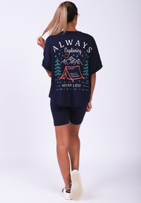 T-shirt surdimensionné bleu marine avec un graphisme de camping, associé à un short sombre et des baskets blanches. Coupe décontractée, manches courtes et ourlet arrondi.