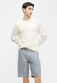 Νέος άνδρας στέκεται με τα χέρια πίσω από την πλάτη, φορώντας πολλαπλά στρώματα λευκών μακρυμάνικων και κοντομάνικων πουκαμίσων και μπλε ριγέ σορτς μέχρι το γόνατο.