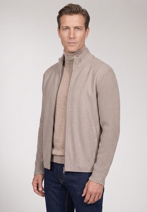 Mann mit hellbraunen Haaren, der eine beige Jacke mit Reißverschluss über einem beigen Rollkragenpullover und dunkelblauen Jeans trägt, steht vor einem einfarbigen Hintergrund.