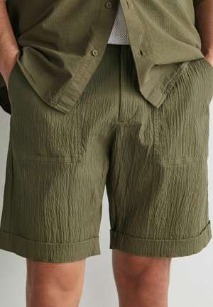 Homme portant un short texturé vert olive et une chemise ouverte assortie, les mains dans les poches, montrant les détails du tissu et un style décontracté.