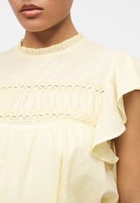 Femme portant un chemisier jaune clair avec col à volants, détails en dentelle et plis, et manches flottantes sur fond blanc.