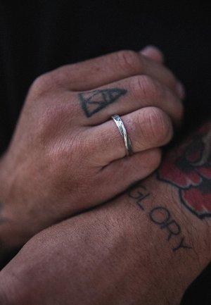 Silver ring with a textured surface worn on a hand with tattoos. Hands clasped, showing details of skin texture and ink patterns.