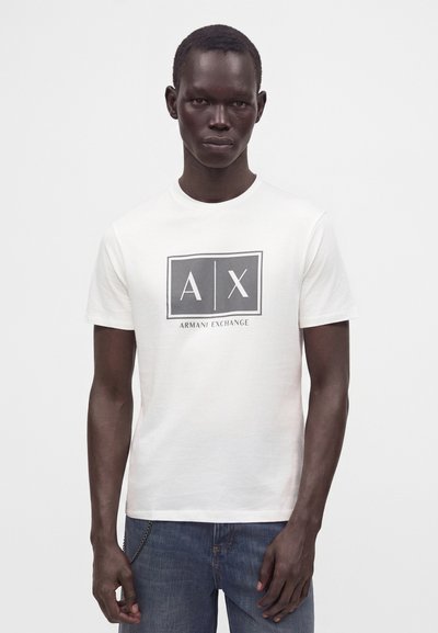 Млад мъж с тъмна кожа, носещ бяла тениска Armani Exchange и сини дънки, стоящ срещу светъл плътен фон.