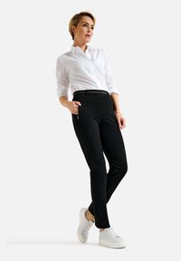 Zwarte op maat gemaakte broek met zijzakken met rits, gecombineerd met een witte blouse en witte sneakers, staande tegen een neutrale achtergrond.