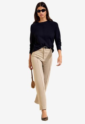 Femme marchant portant un haut noir à manches longues, un pantalon beige, des talons noirs, des lunettes de soleil et tenant un petit sac à main marron.