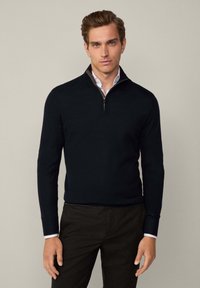 Mörkblå kvartsdra sweater i mjukt stickat material, med ståkrage och långa ärmar, kombinerad med mörka byxor.