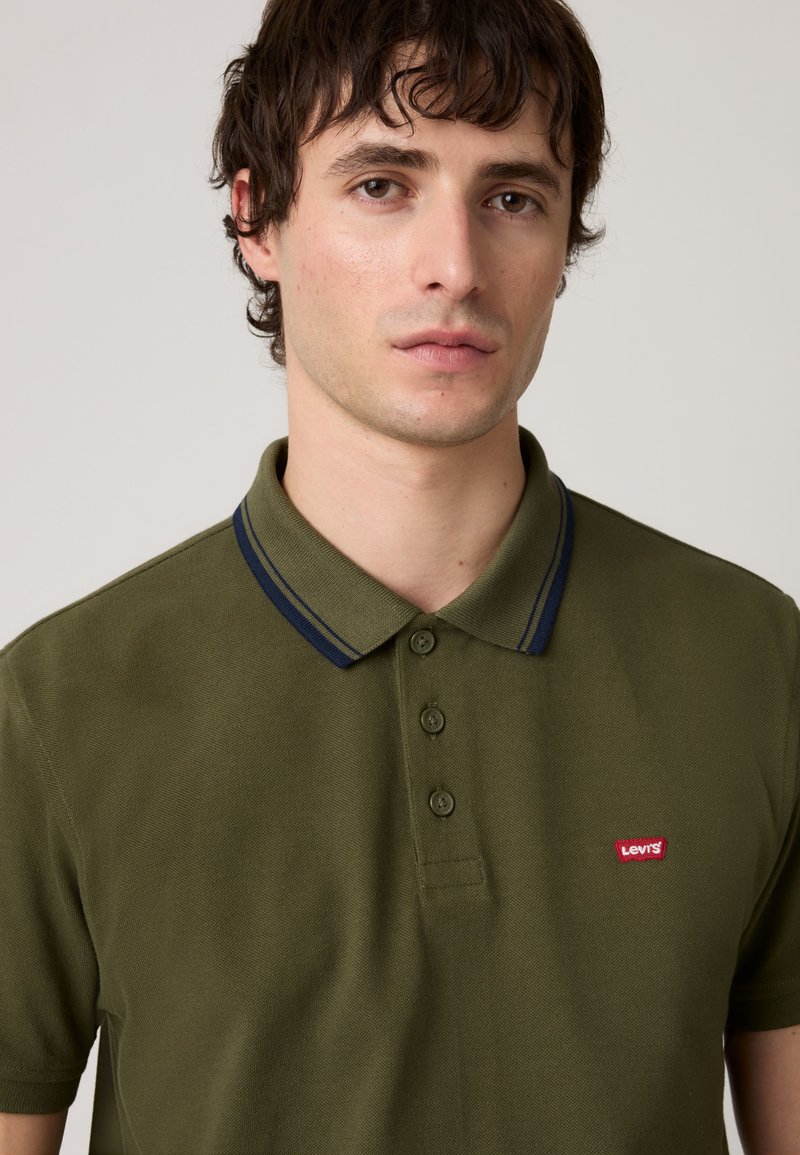 Jeune homme aux cheveux courts et foncés portant un polo vert olive avec bordure bleu marine et un logo Levi's rouge sur la poitrine.