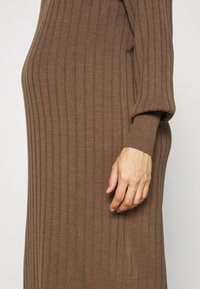 Vista laterale di una persona che indossa un vestito a maglia a coste marrone con maniche lunghe, con la mano rilassata al suo fianco su uno sfondo neutro.