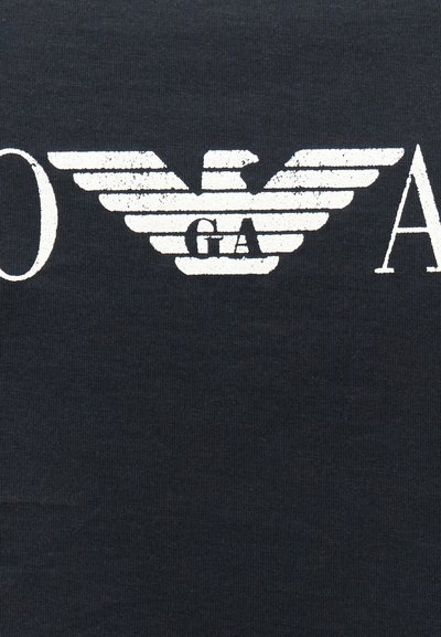 Λευκό στυλιζαρισμένο λογότυπο αετού με οριζόντιες ρίγες και τα γράμματα "G A" στο κέντρο σε σκούρο υφασμάτινο φόντο.